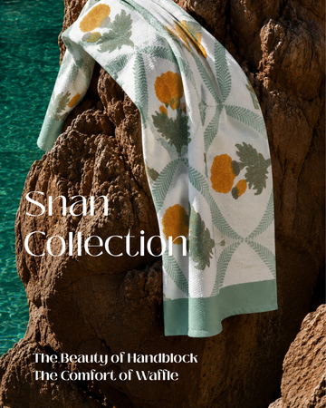 A patterned handblock-printed towel draped over a rocky shoreline with turquoise water in the background. The towel has a white base with sage green geometric lattice patterns and mustard yellow floral motifs.